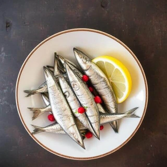 A can of sardines with a forkful on a plate, surrounded by fresh herbs and lemon slices.