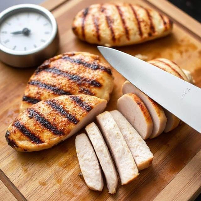 Grilled chicken resting on a wooden cutting board, being sliced into juicy strips against the grain with a sharp knife